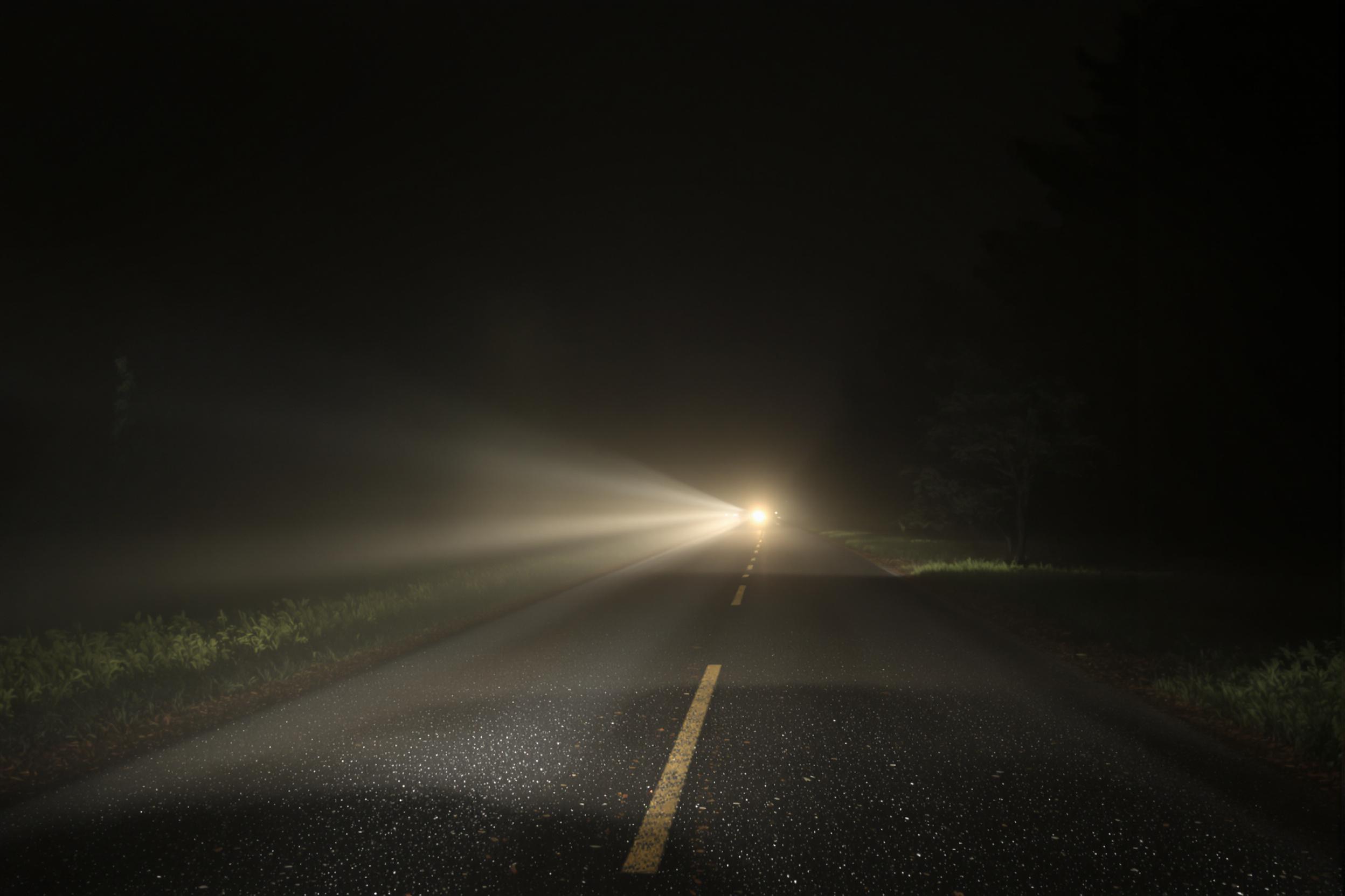 Car headlights pierce fog on rural road at night